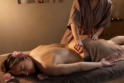 Person receiving back massage in spa. Therapist using wooden tool. Beige setting, floral accents.