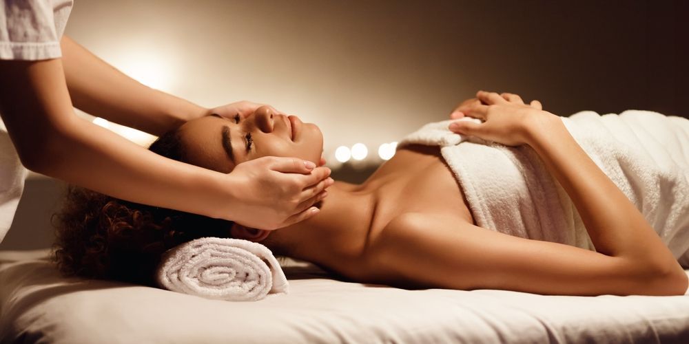 Person receiving a face massage while lying on a massage table, wrapped in a white towel.