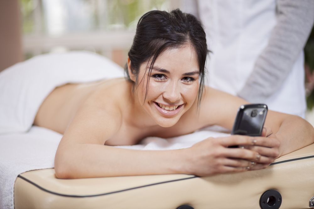Woman on massage table smiles at camera while using a phone, spa setting.