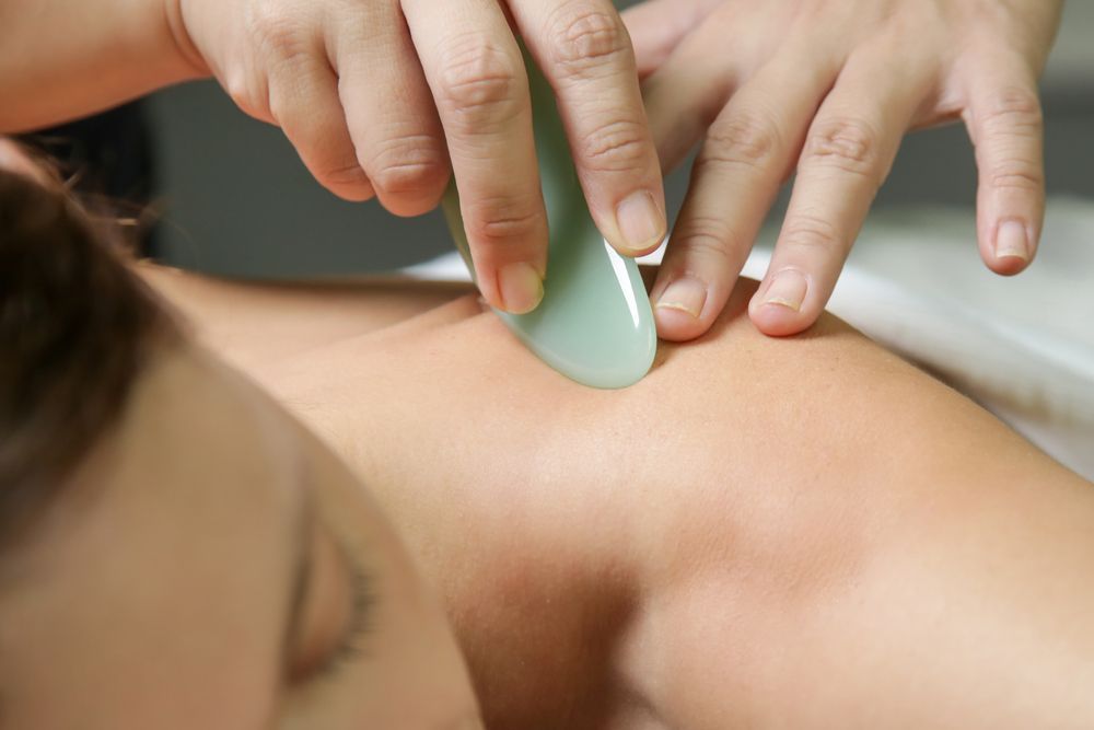 Person receiving gua sha treatment on their shoulder. A hand holds a jade tool.