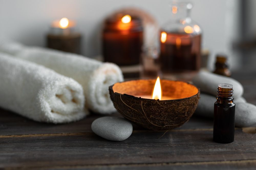 Relaxing spa setup: rolled white towels, lit candles, essential oil bottle, and river stones on wood.