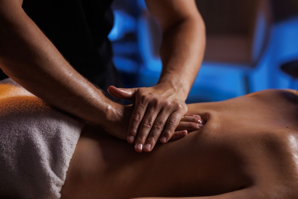Hands giving a back massage; person lying face down, torso exposed, on a massage table, in a dim spa setting.