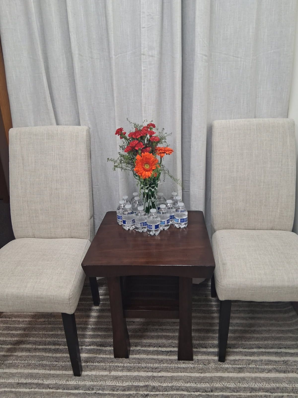 Two beige chairs flanking a dark wood table with a vase of orange and red flowers, set against a light curtain.