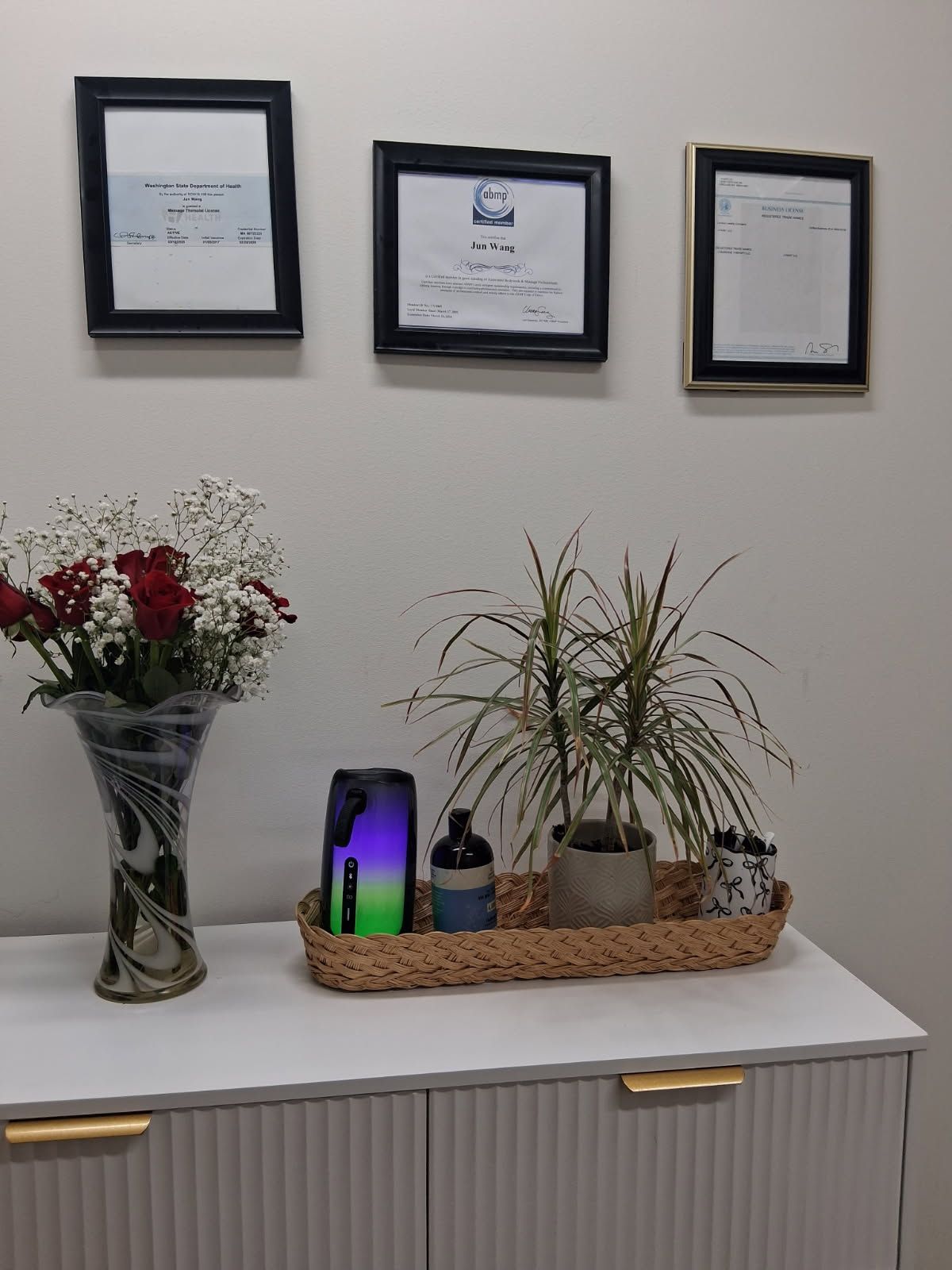 A white cabinet with decor, including flowers, plants, and framed certificates on the wall.