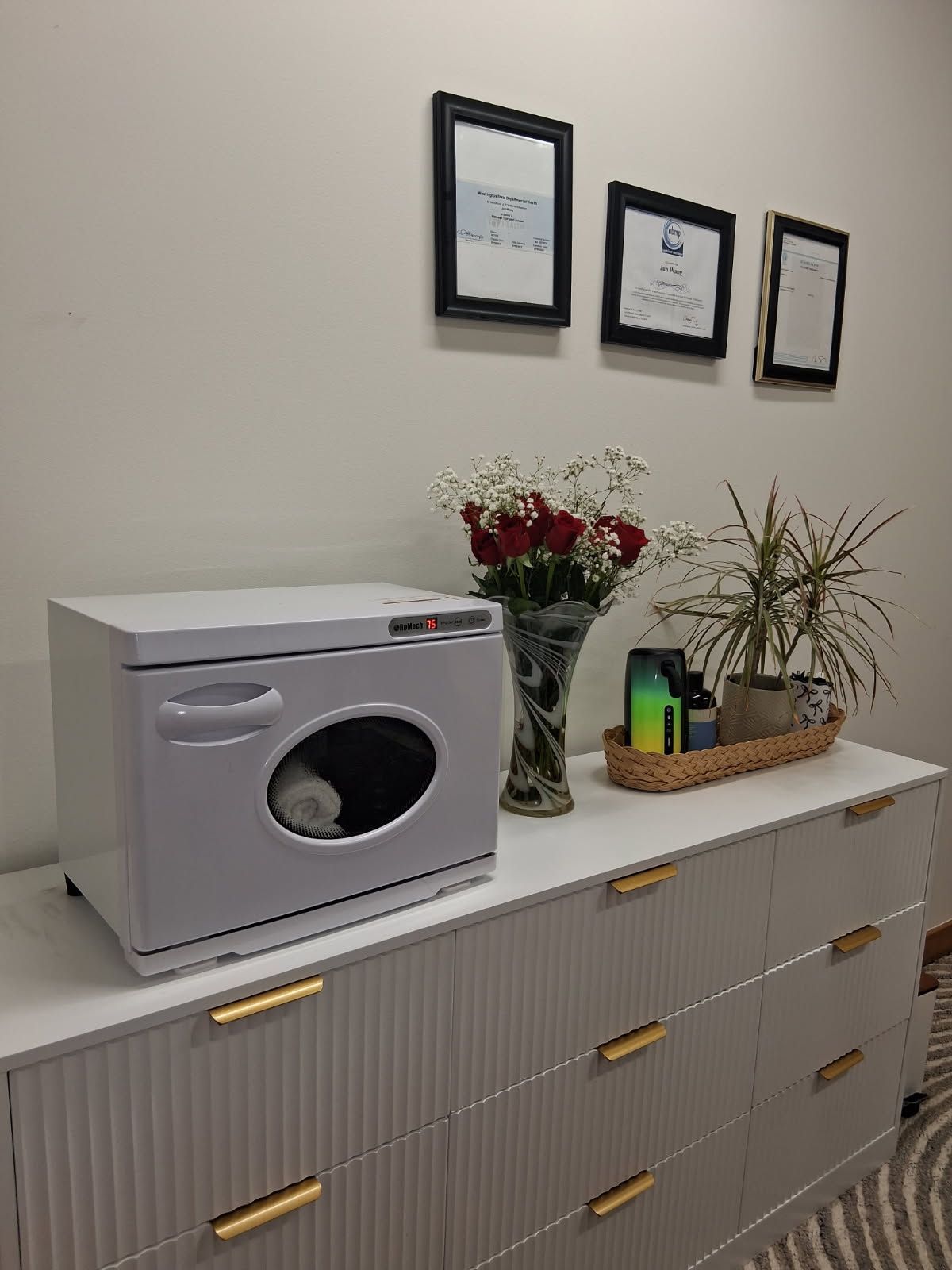 White cabinet with flowers, plants, and a towel warmer. Art on the wall above.