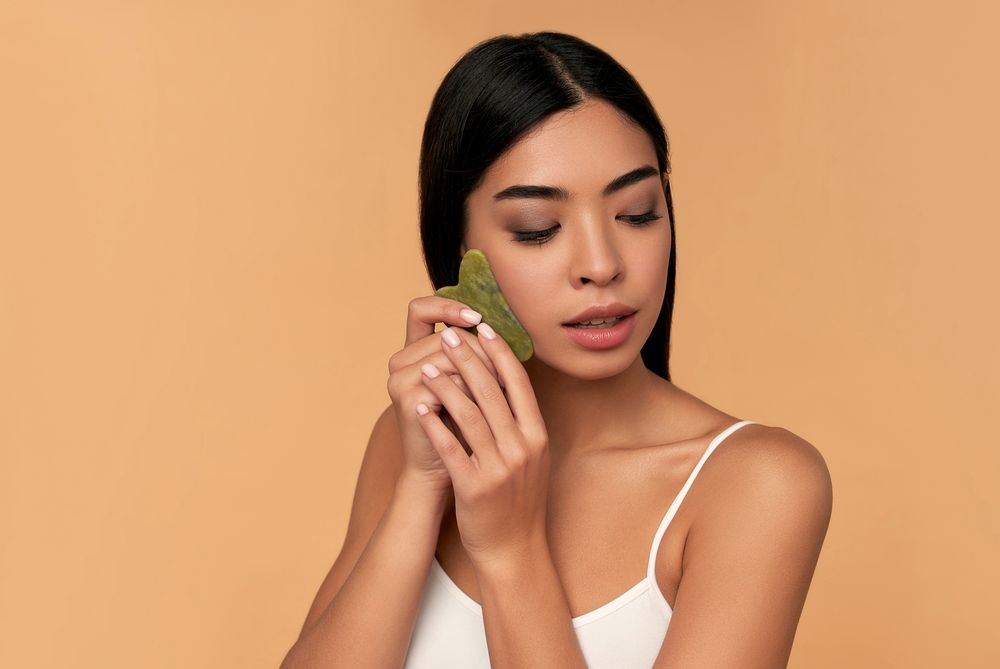 Woman using a jade gua sha tool on her face, in front of a neutral background.