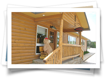 A teddy bear is sitting on the porch of a log cabin.