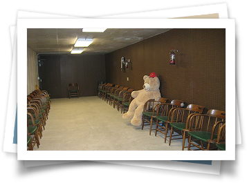 A large teddy bear is sitting in a room with chairs.