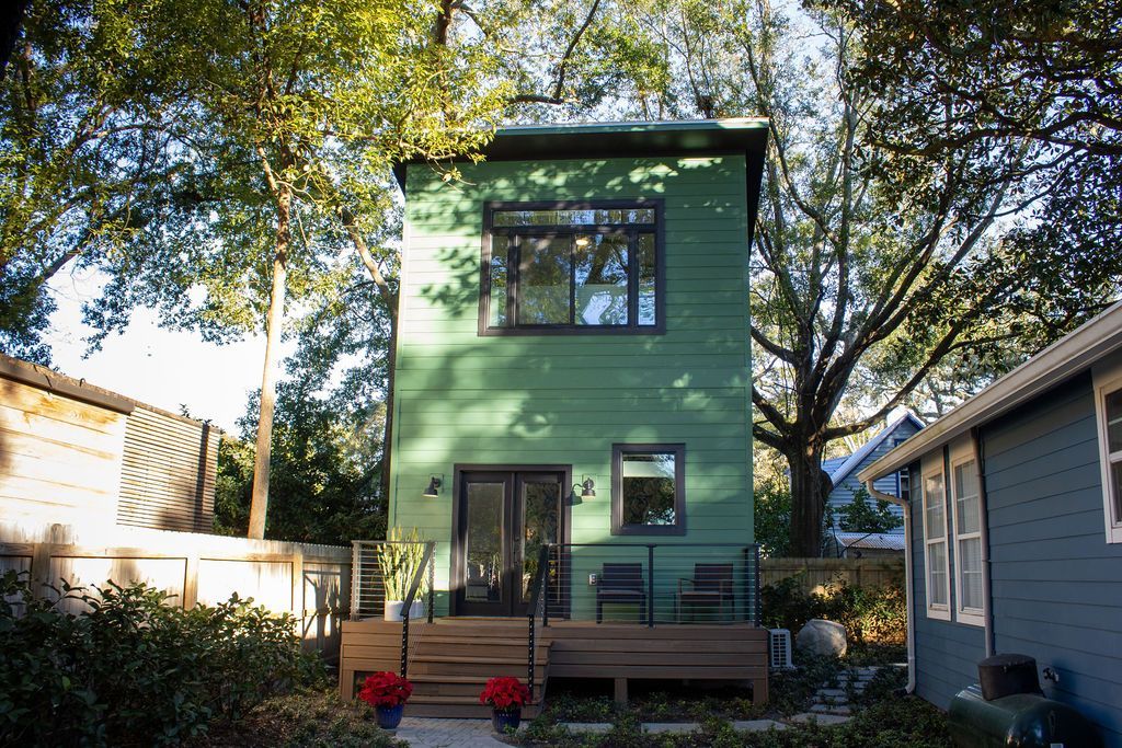 Green two-story tiny house with a deck, surrounded by trees and other small houses in a yard.