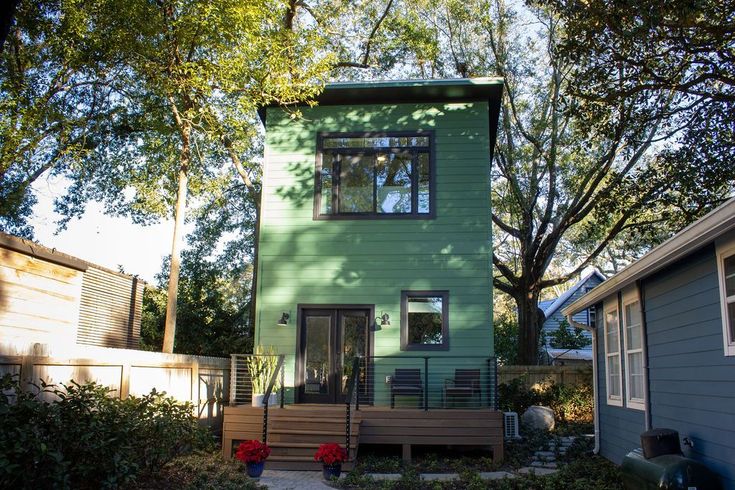 Green two-story tiny house with a deck, surrounded by trees and other small houses in a yard.