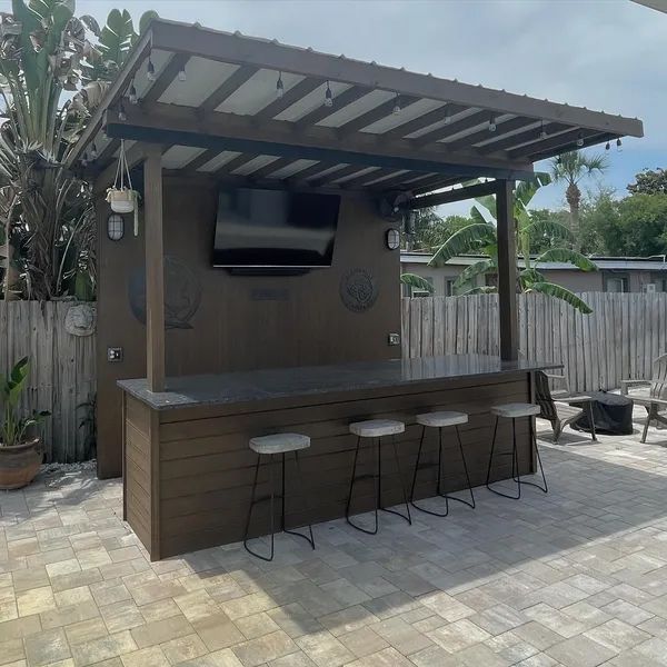 Outdoor bar with brown wood siding, stools, and a TV, on a brick patio.