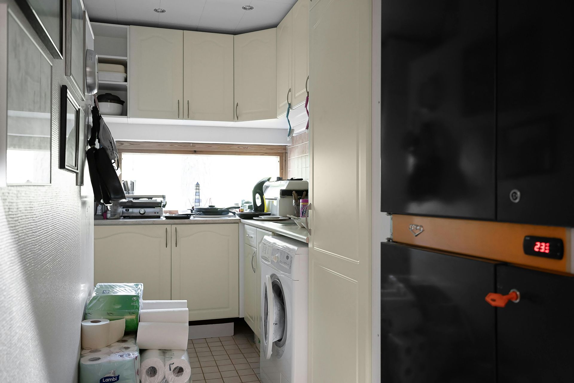 Small kitchen with off-white cabinets, a window, and a washing machine. Toilet paper rolls and black cabinets are also present.