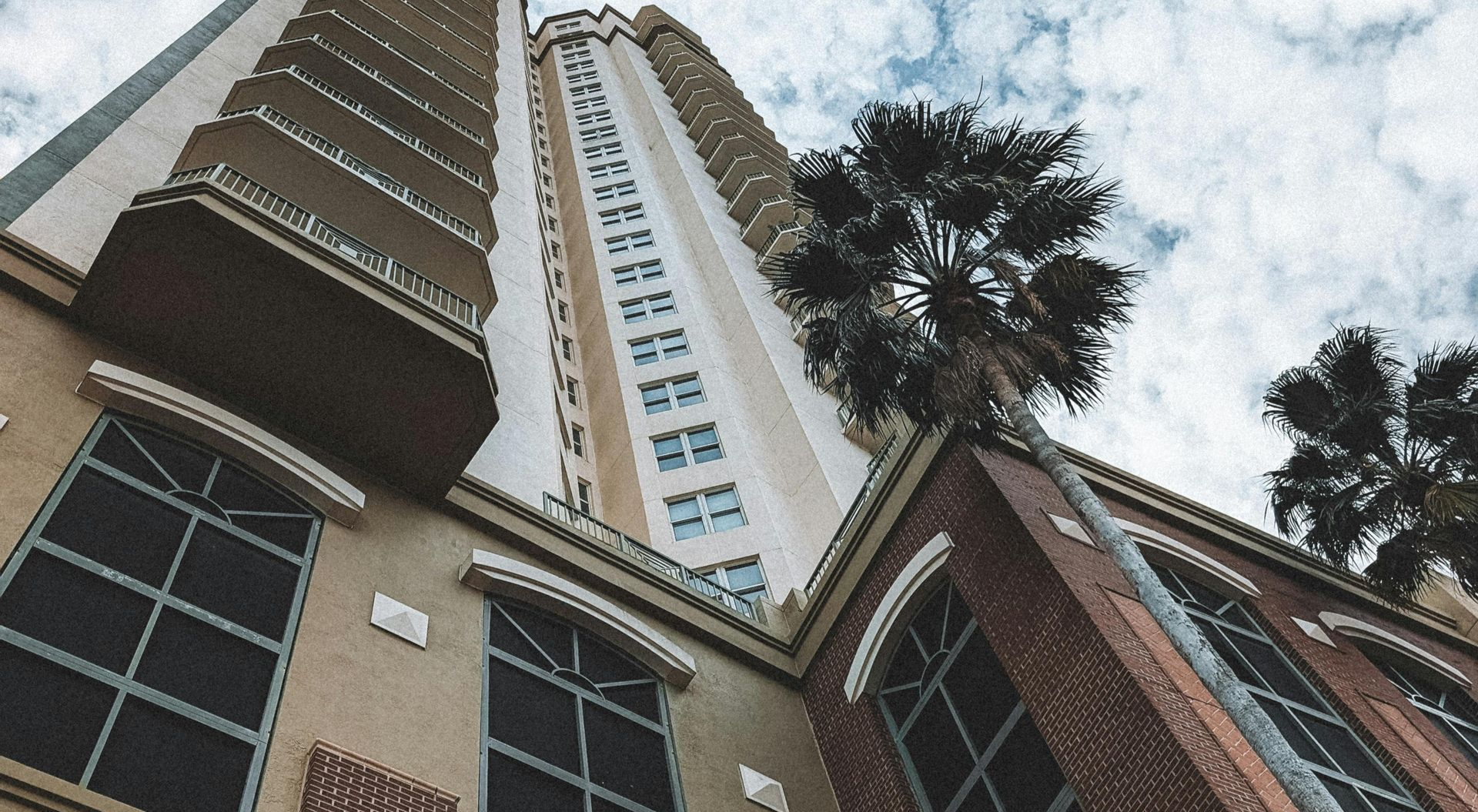 Tall beige and brown building with arched windows, balconies, and palm trees. Cloudy sky.