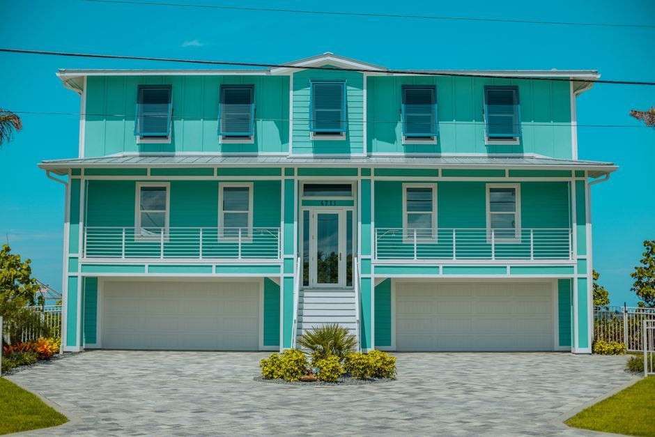 Turquoise two-story house with white trim, balconies, and two garage doors under a clear sky