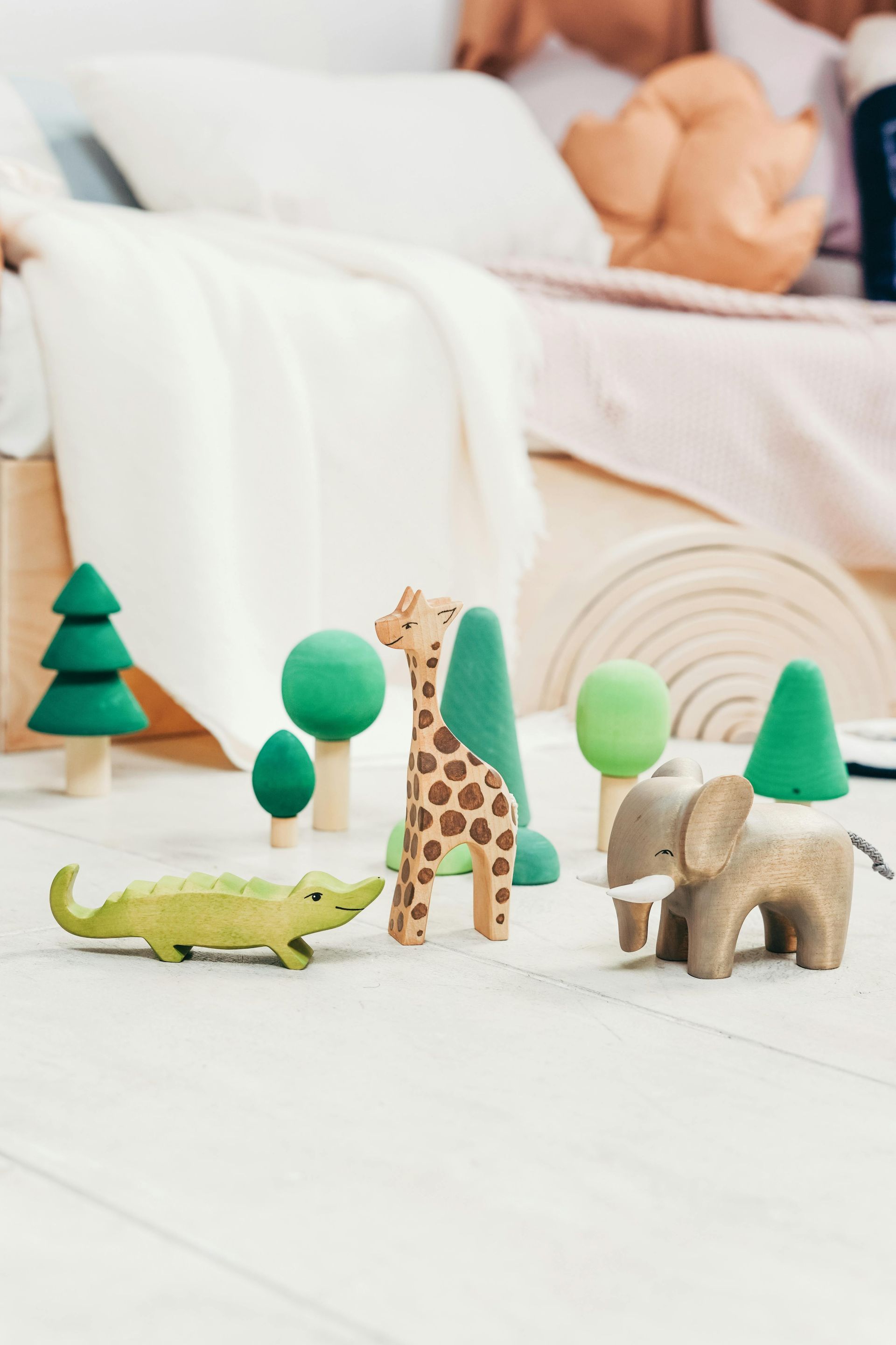 Wooden animal toys (crocodile, giraffe, elephant) and trees on a white floor in front of a bed.