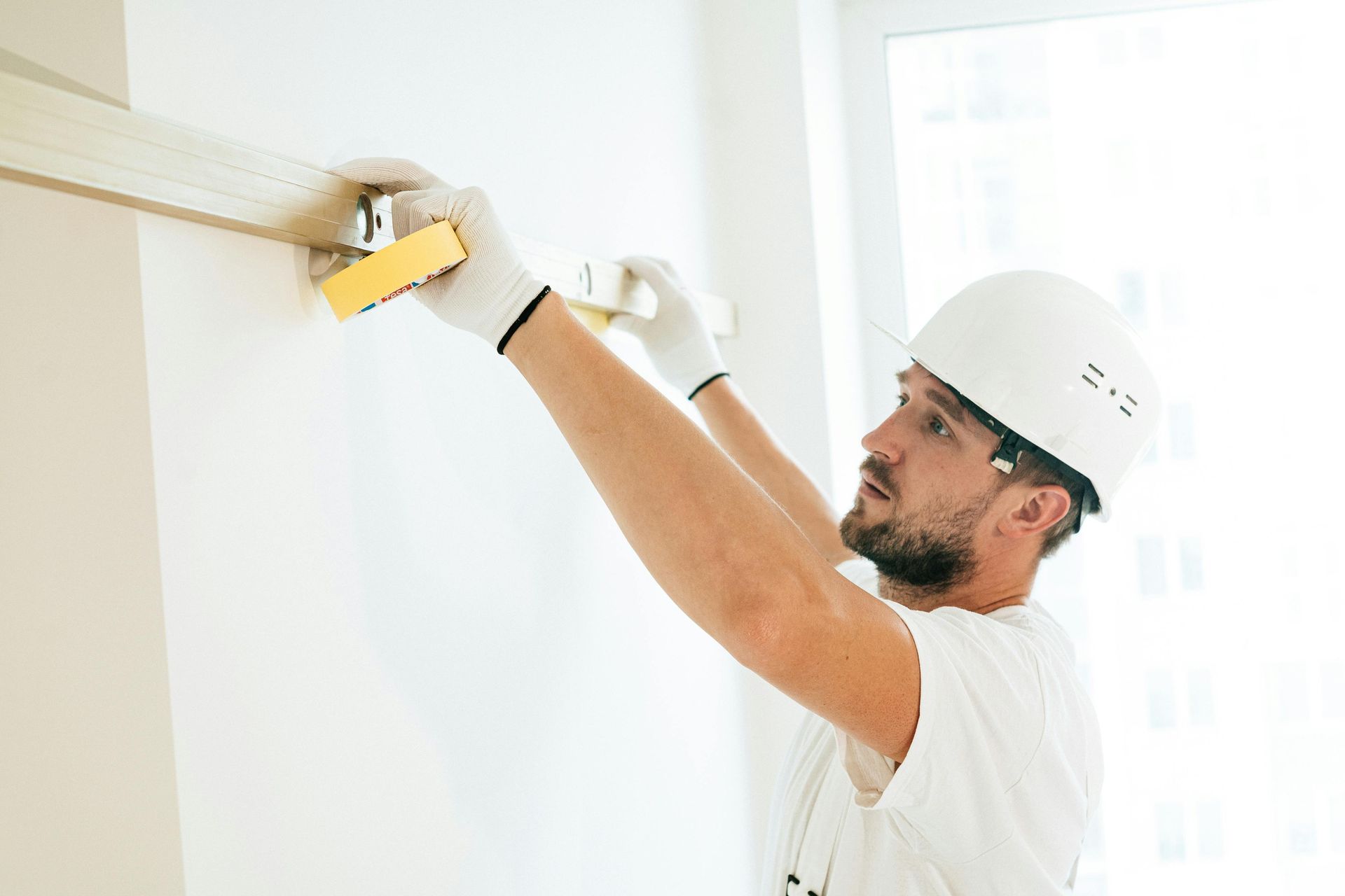 Person wearing hard hat and gloves installing molding on a white wall near a window.
