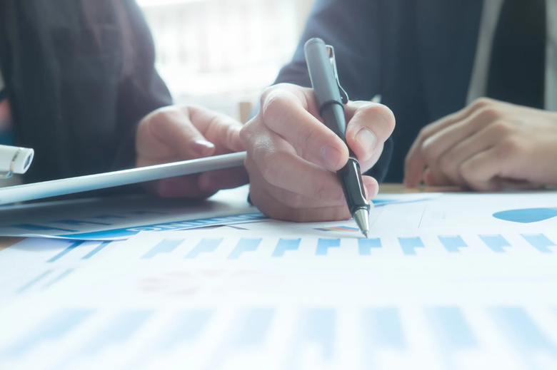 Person's hand writing on a document with charts, alongside another person; office setting.