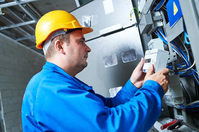 Electrician in blue uniform and hard hat working on electrical panel.