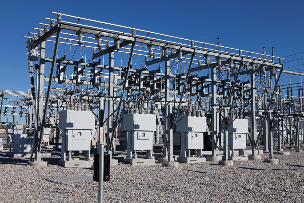 Electrical substation with metal structures, white boxes, and wires against a blue sky.