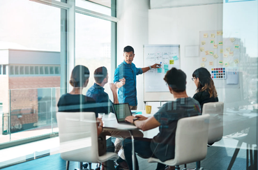 Team in meeting; man points at whiteboard with sticky notes, others look on. Bright, modern office space.