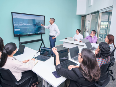 A man points at a screen in a bright office. People sit at desks with laptops, listening.