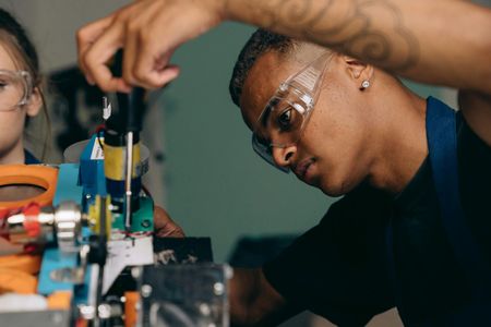Man in safety glasses uses a screwdriver on machinery; a person is partially visible on the left.
