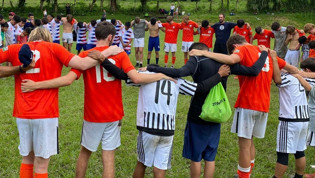 Two soccer teams with arms around one another pray together.