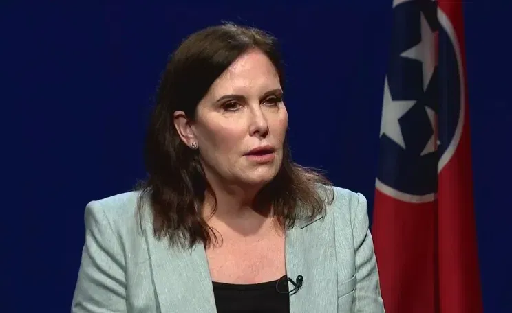 A woman in a suit is sitting in front of a flag.