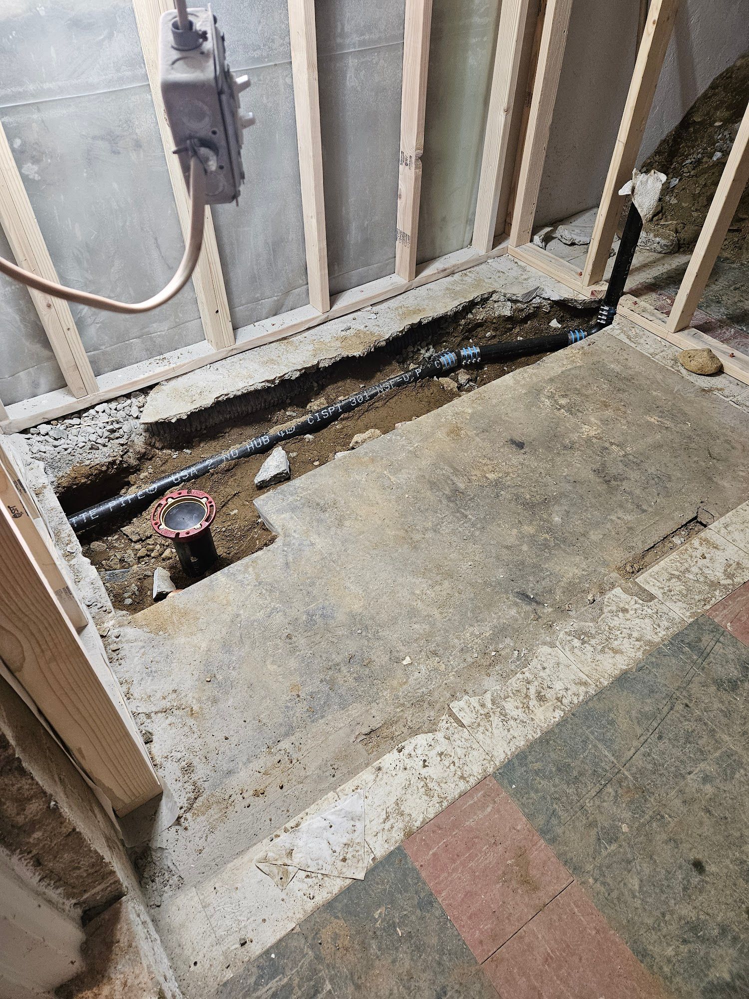 A basement floor cut out to reveal exposed plumbing pipes and a drain connection between wall studs.