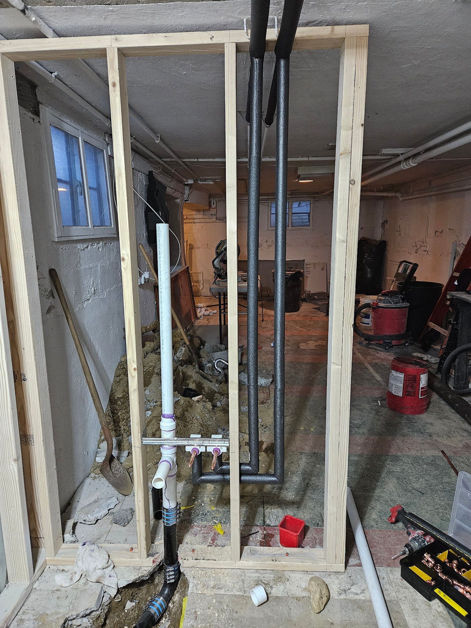 Wooden wall frame in a basement construction site with exposed vertical plumbing pipes and unfinished debris.