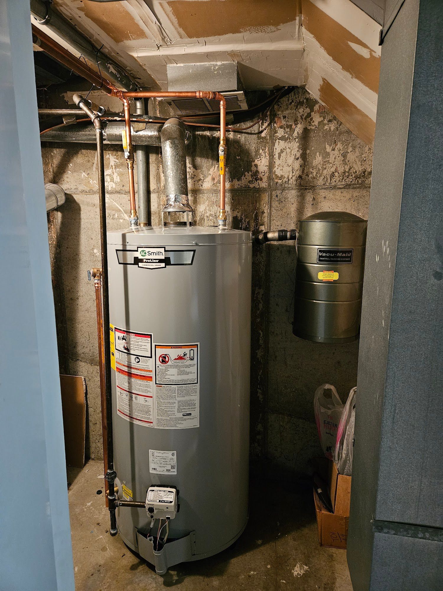 A grey water heater with copper piping and an expansion tank in a basement against a concrete block wall.