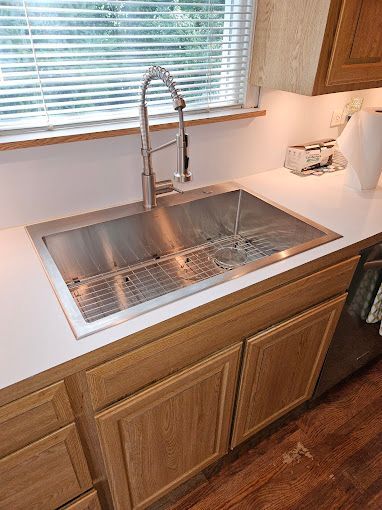 A stainless steel kitchen sink with a high-arch faucet, set in white countertops above wooden cabinets near a window.