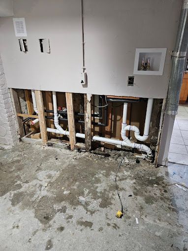 An open section of a basement wall revealing plumbing pipes and electrical wiring behind a concrete floor.