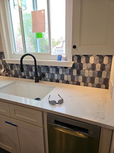 A modern kitchen sink area with white countertops, a dark faucet, and a grey and white elongated hexagonal tile backsplash.
