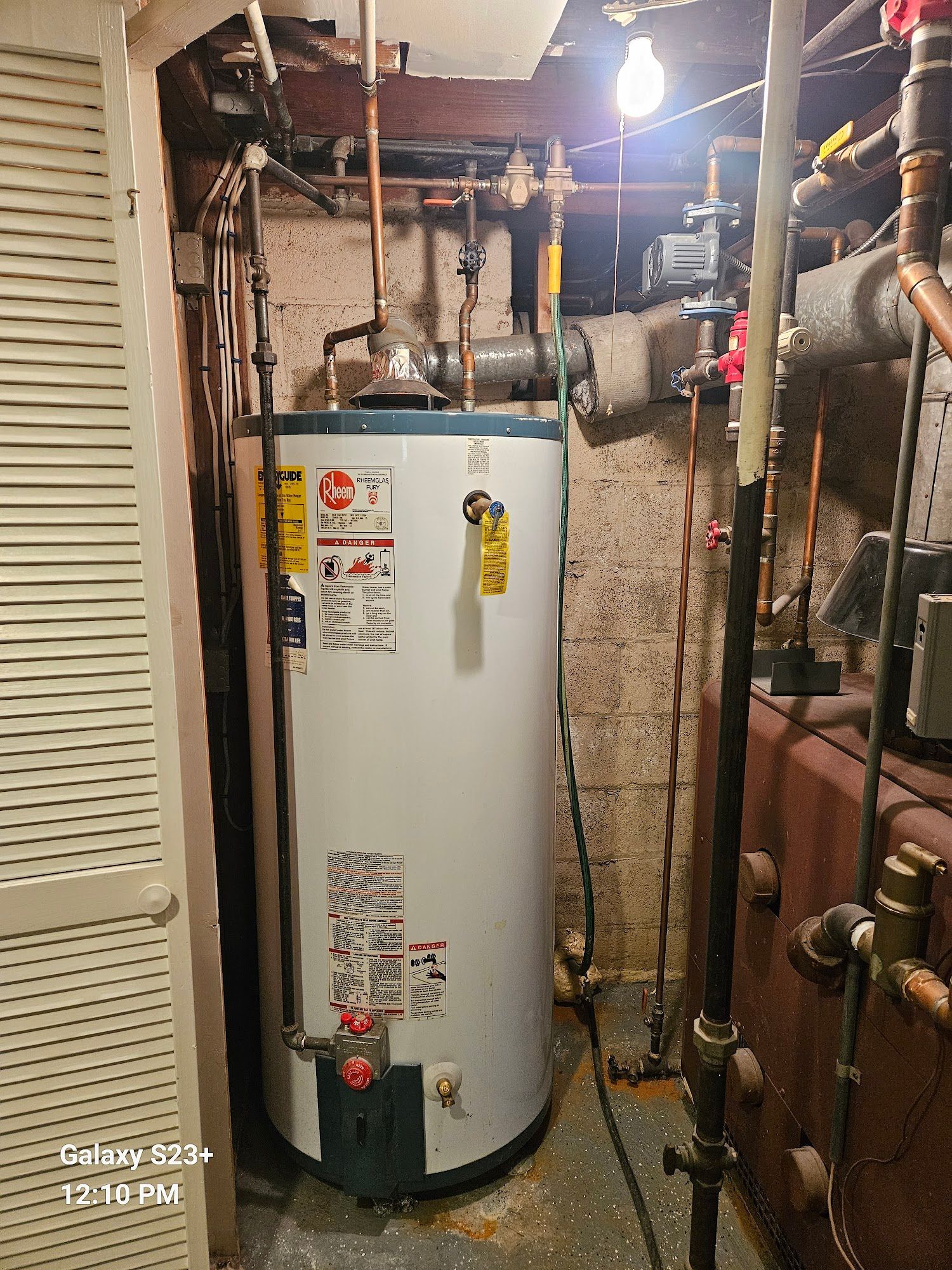 A white water heater stands in a basement next to copper pipes and a brown industrial furnace.