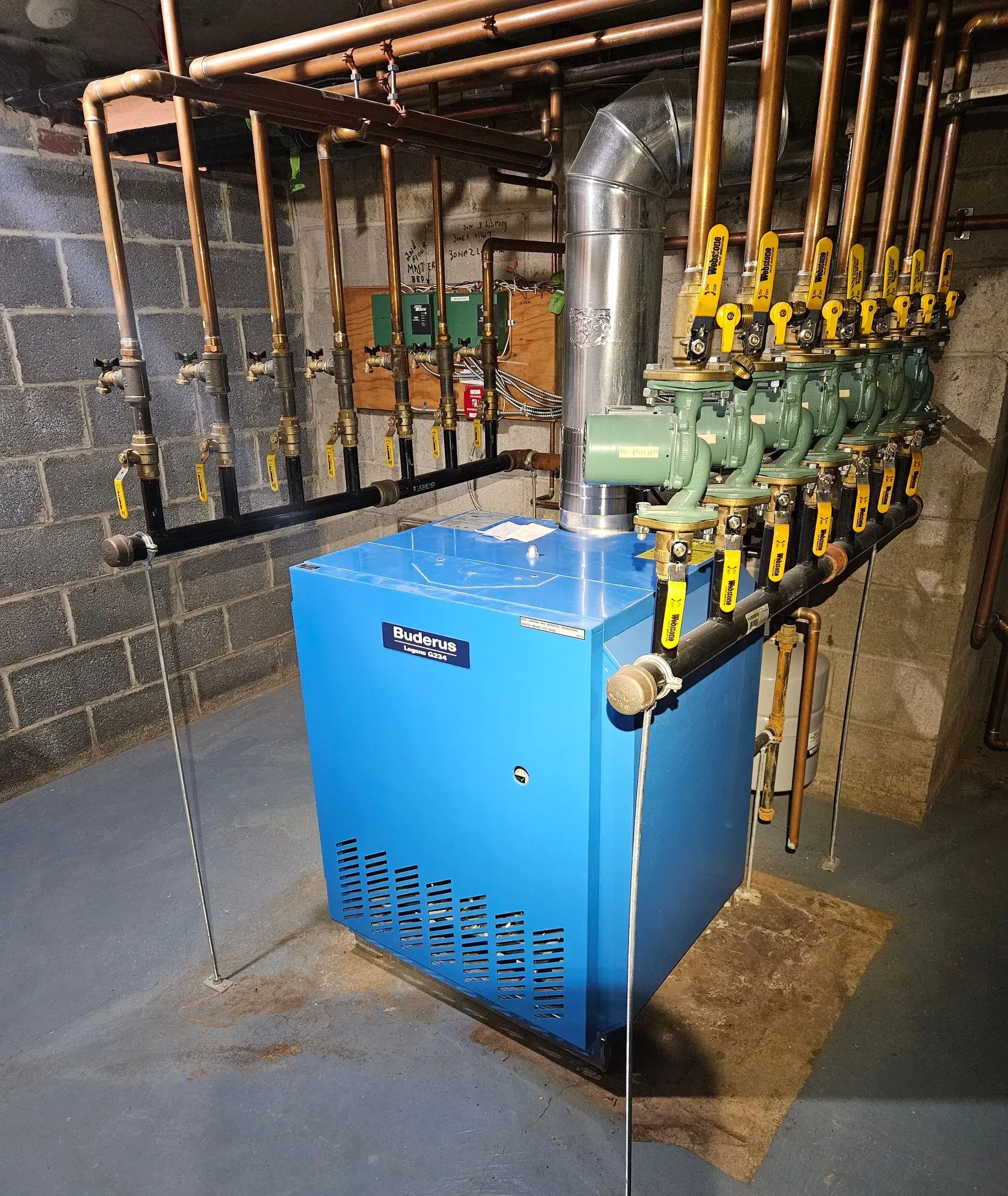 A bright blue residential heating boiler with copper piping and multiple green pumps mounted in a basement.