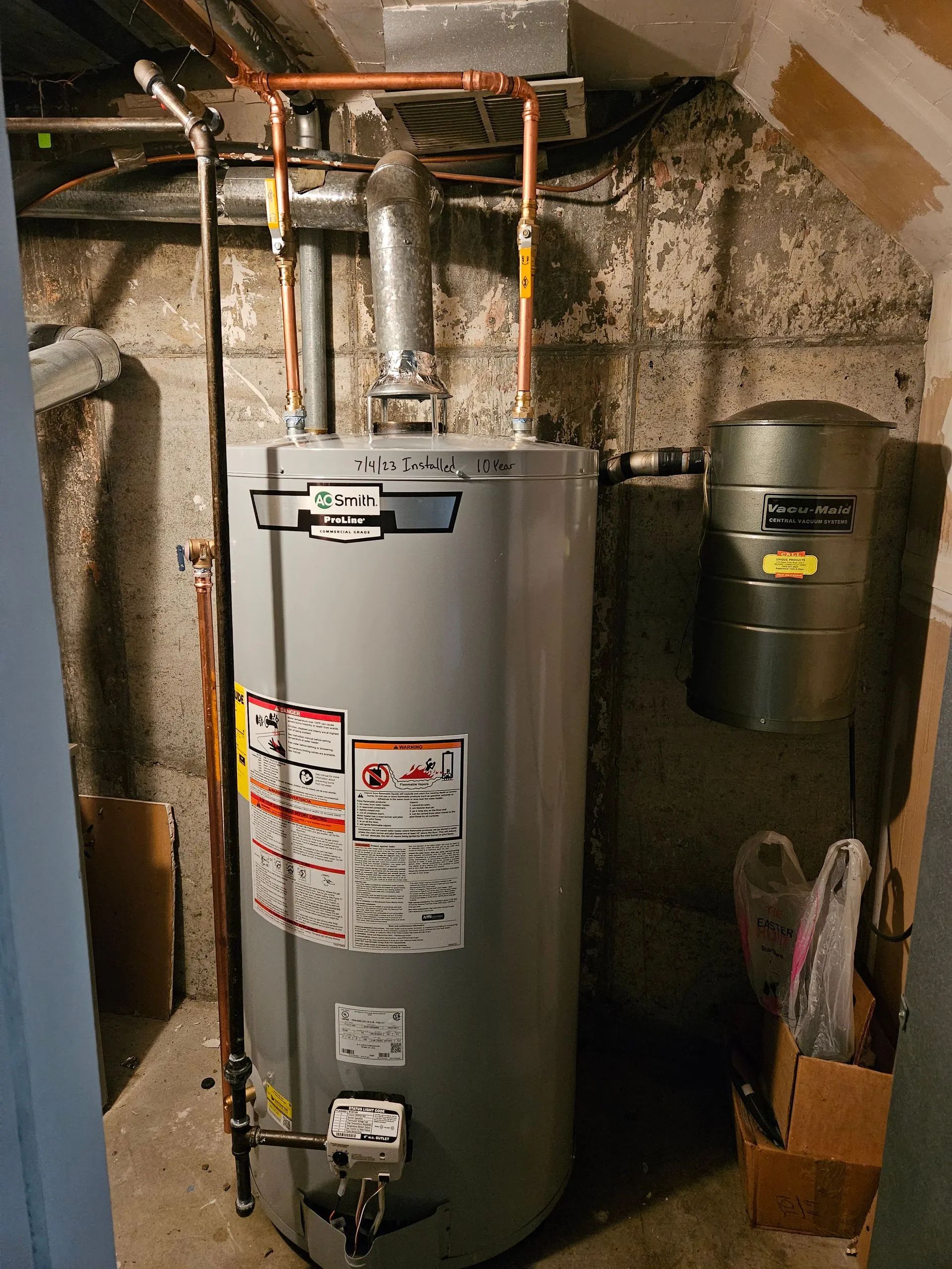 A grey Rheem water heater in a basement, connected to copper piping and a metal expansion tank against a concrete wall.