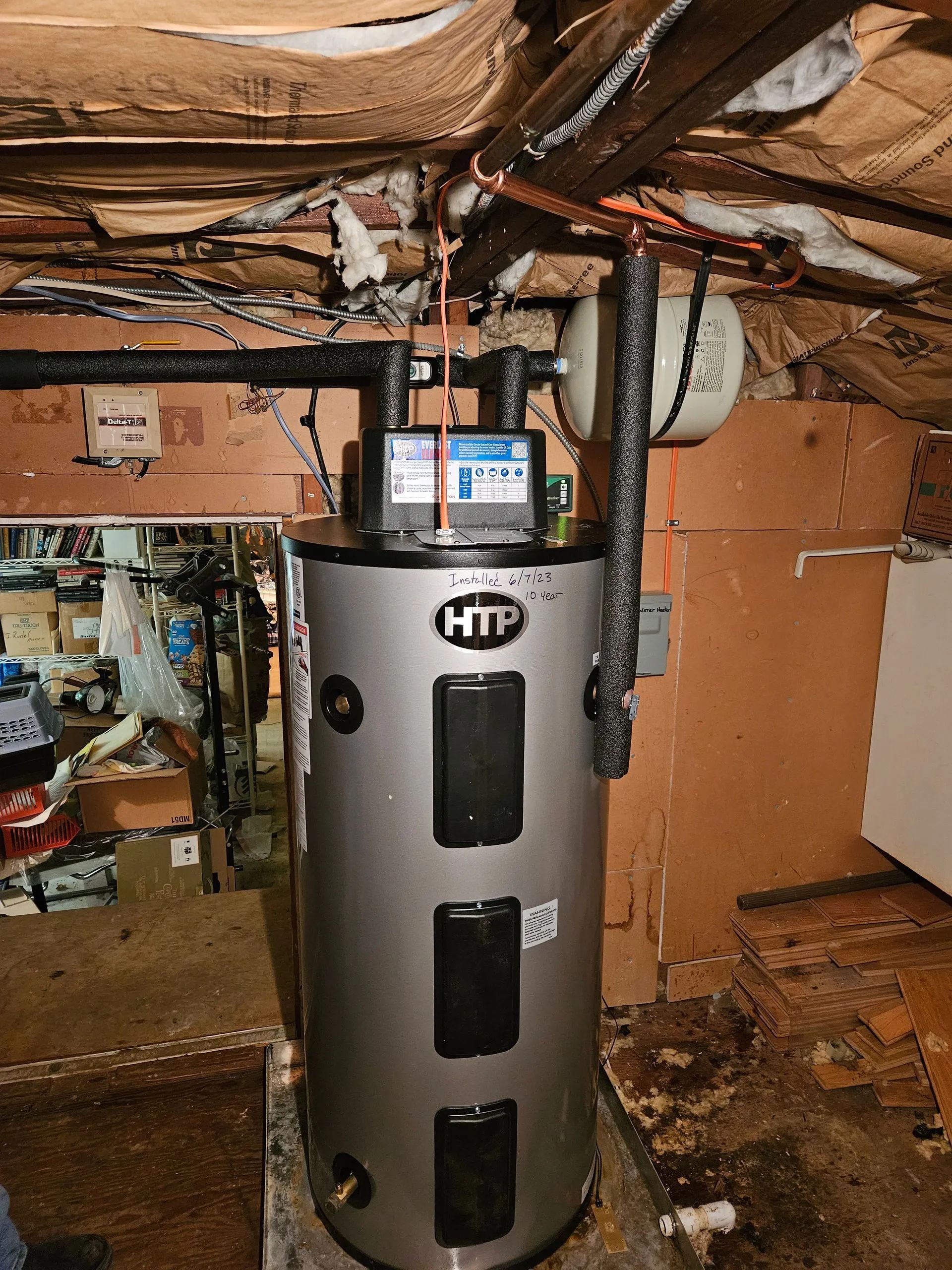 A grey HTP water heater stands in a basement utility area, connected to insulated pipes and a white expansion tank above.