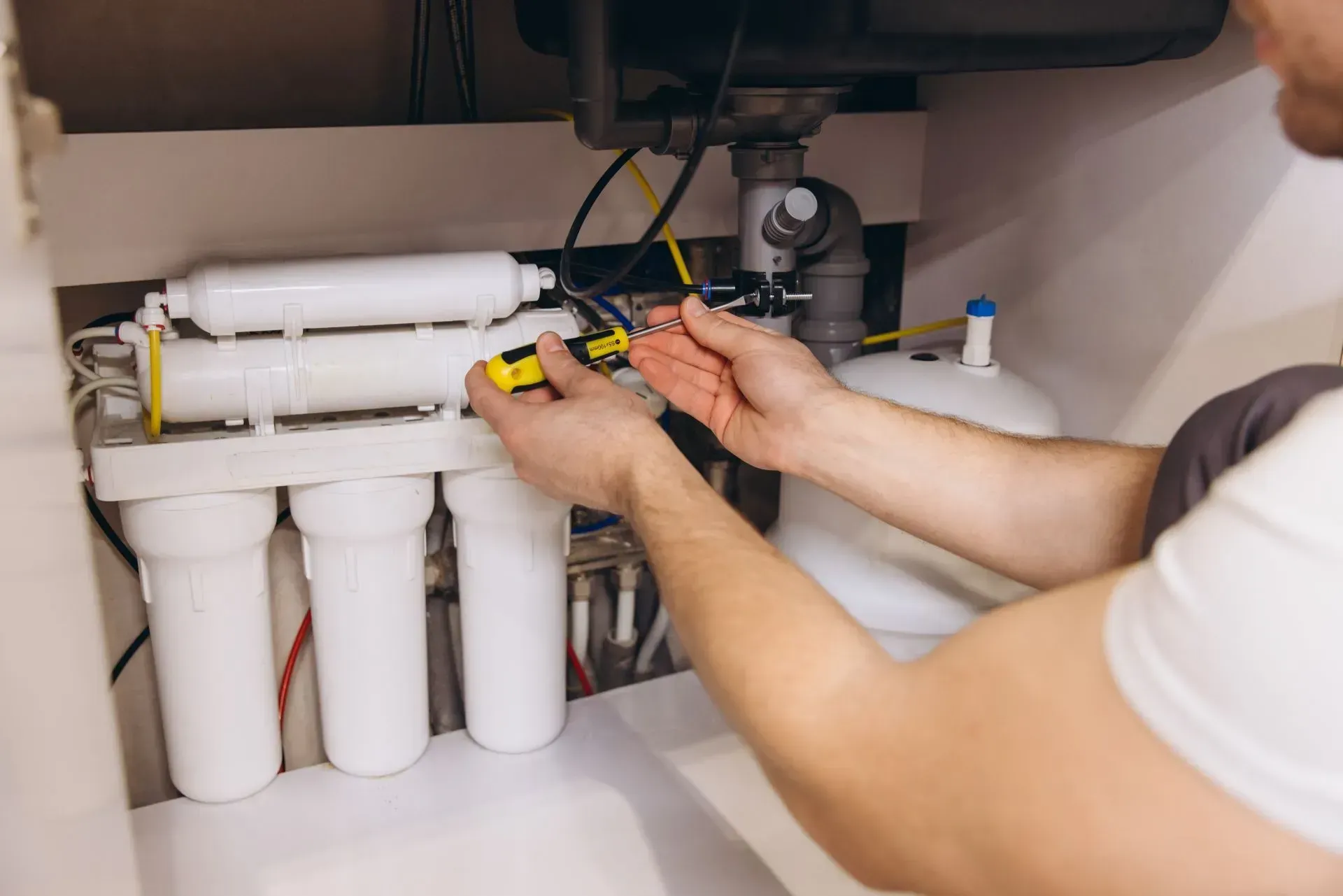 A person uses a screwdriver to repair a white reverse osmosis water filtration system installed under a kitchen sink.