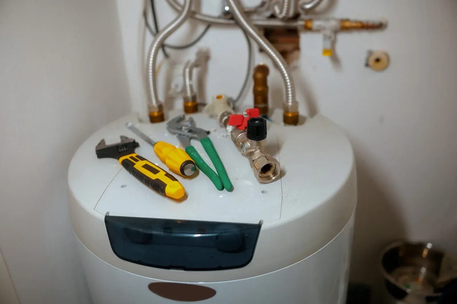 A white water heater with tools, including a wrench, screwdriver, and pliers, and a brass valve resting on top.