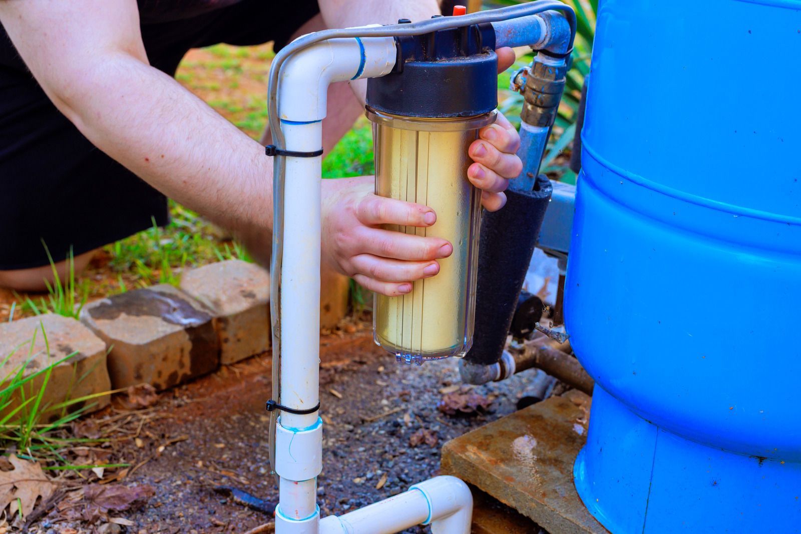 A person holding a clear water filter housing attached to blue pipes and a blue barrel outdoors.