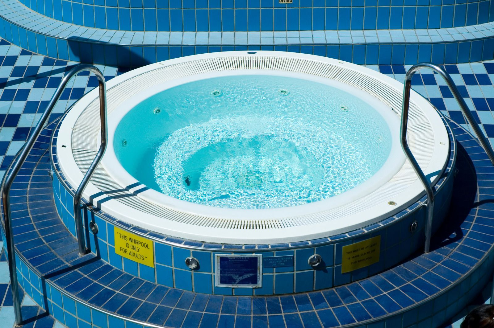 A round, blue-tiled hot tub filled with clear water, featuring metal handrails and yellow safety signs on its exterior.