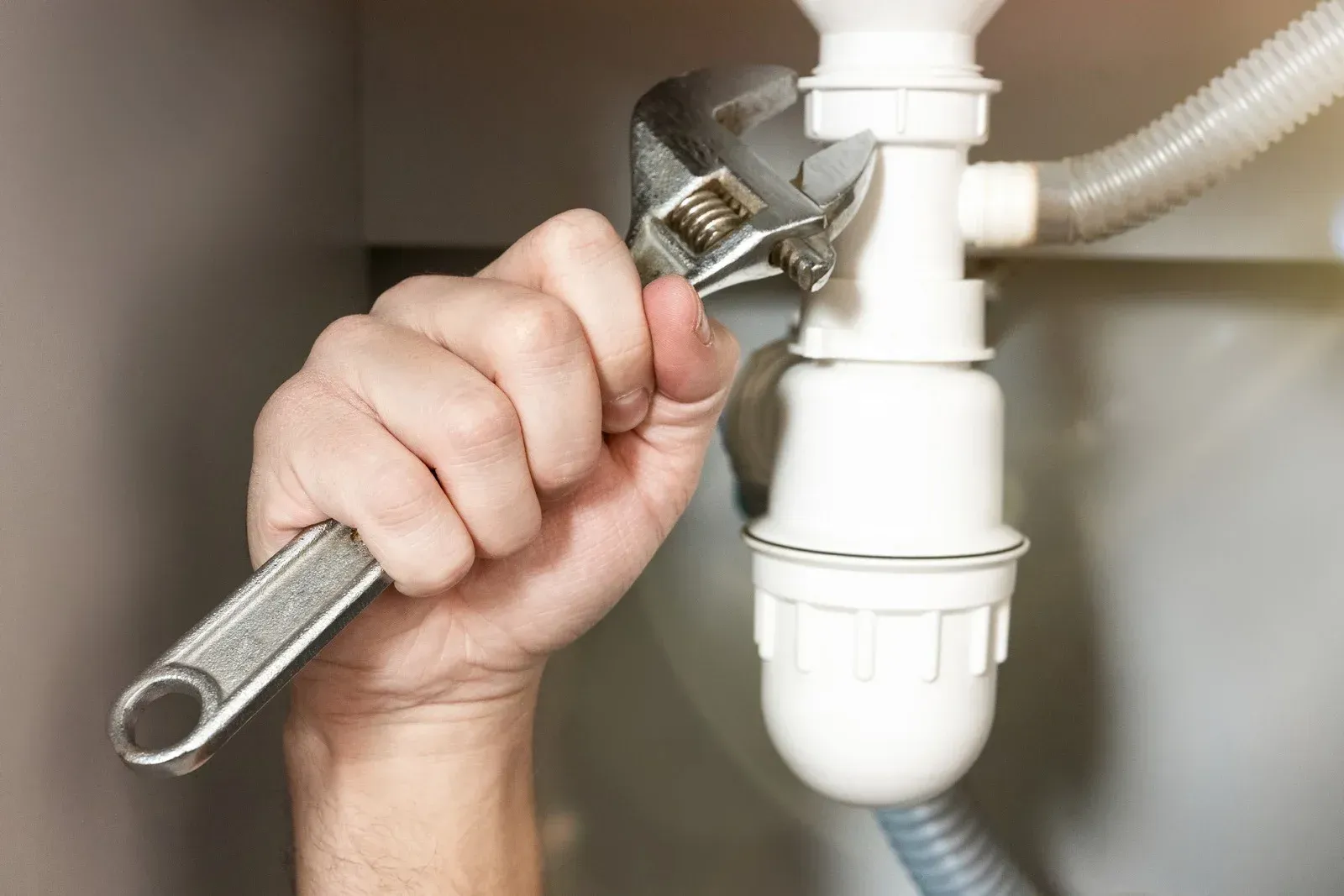 A hand using an adjustable wrench to tighten a white plastic sink pipe fitting.