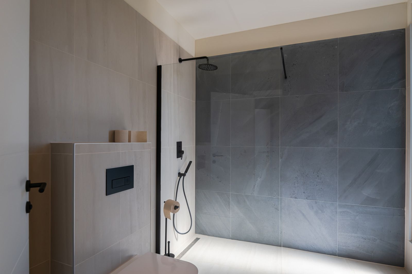 A modern bathroom with a charcoal tiled walk-in shower, a black glass screen, and a wall-mounted toilet.