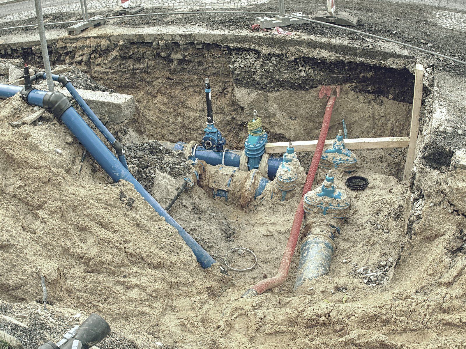 A deep excavation in dirt exposing blue water utility pipes, valves, and a red pipe segment.