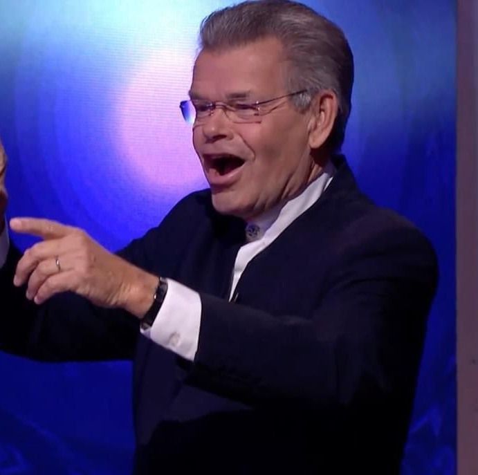 Man met lang krullend haar, gekleed in een blazer, sjaal en spijkerbroek, staat voor een muur van beton en hout.
