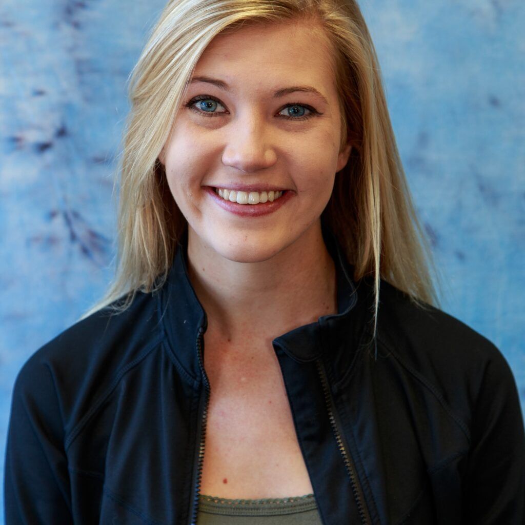 Blond woman smiling, wearing black jacket and olive green top, blue background.