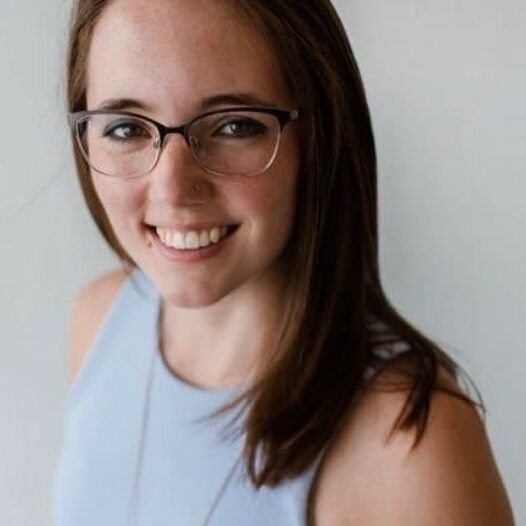 Woman with glasses smiles, wearing a light blue top, against a light grey background.