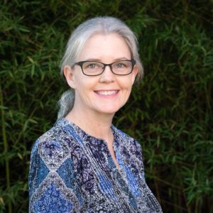 Woman with glasses smiling, standing outdoors in front of green bamboo, wearing a blue patterned shirt.