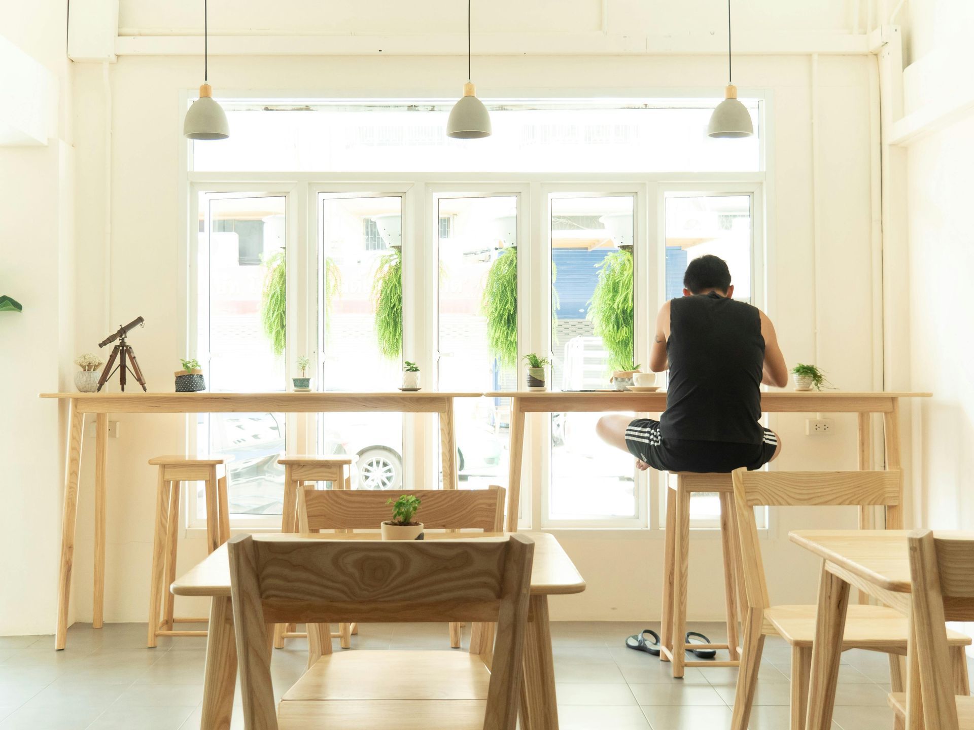Man sitting at a wooden counter in a bright cafe, facing a window. Other chairs and tables are nearby.